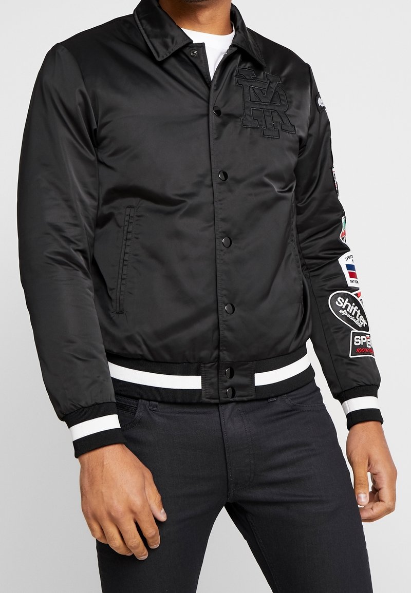 Černá bomber jacket z saténového materiálu, žebrované manžety a lem s bílými pruhy, vyšívané logo na hrudi a různé nášivky na rukávech.