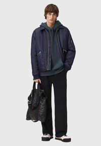 Námořnická bombardérka s dvěma zipovými kapsami, přes kterou je oblečená zelená mikina s kapucí. Model drží prošívanou černou tašku; černé kalhoty a boty dokončují vzhled.