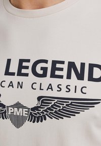 Bež majica s okruglim izrezom, s velikim crnim tekstom koji glasi "LEGEND" i amblemom s krilima i trakama.