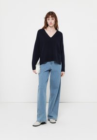 Maglione a V collo in maglia di colore blu navy, con maniche lunghe, abbinato a jeans larghi di colore blu chiaro e stivali grigi, completato da una piccola borsa bianca.