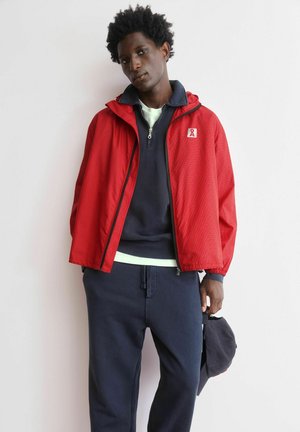 Man draagt een rode jas met ritssluiting over een marineblauwe half-rits trui en marineblauwe broek, houdt een donkere honkbalpet naast zich vast.