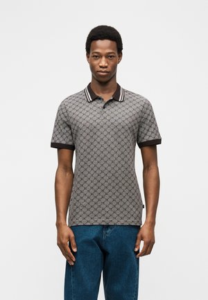 Homme debout, faisant face, portant un polo gris à motifs avec col et poignets foncés, et un jean bleu, sur un fond blanc.