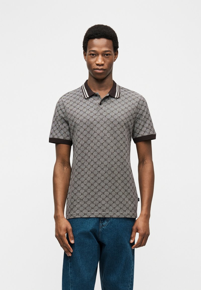 Άνδρας που στέκεται με το πρόσωπο προς τα εμπρός, φορώντας γκρι πόλο με σχέδια, σκούρο γιακά και μανσέτες, και μπλε τζιν, σε λευκό φόντο.