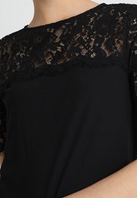 Top negro con un yugo de encaje con patrones florales y bordes en forma de escallola, mangas cortas y un cuerpo de tela suave.