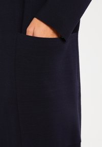 Marineblaues Strickkleid mit langen Ärmeln, seitlicher rechteckiger Tasche, strukturiertem Stoff und lockerer Schnitt.