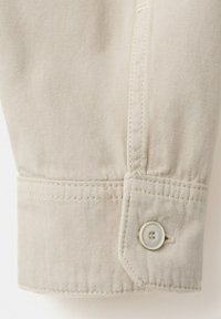 Gros plan sur un poignet de manche en tissu beige avec un seul bouton blanc et des détails de couture visibles.