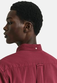 Bordeauxfarbendes Hemd mit hohem Kragen, Knopfdetails und einer Falte im Rücken. Der Stoff wirkt glatt mit einer matten Textur.
