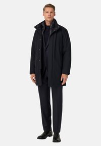 Parka negra impermeable con un cuello alto, botones a presión y un interior acolchado, combinada con pantalones oscuros y zapatos negros.