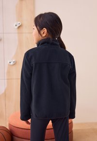 Giacca in pile nera con collo alto, caratterizzata da maniche lunghe e una vestibilità comoda. Tessuto liscio, dettagli cuciti lungo le spalle.