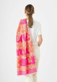 Rosa und orange Strandtuch mit floralem Muster und fettem Text, aus weichem Stoff, mit fransigen Kanten, über die Schulter einer Person gelegt.