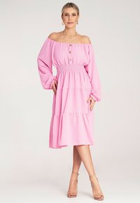 Różowa sukienka na ramiączkach z długimi rękawami, marszczonym pasem, warstwowym krojem, drewnianymi guzikami i miękką teksturą. Noszona z beżowymi szpilkami.