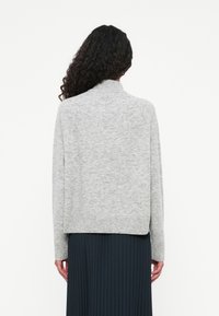 Cardigan de punto gris claro con un cuello alto, dobladillo acanalado y hombros caídos, combinado con una falda oscura plisada. Diseño simple y moderno.
