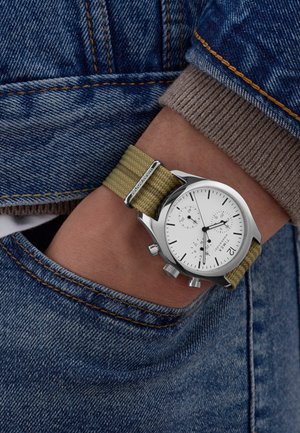 Sølv chronograph ur med hvid urskive og grøn tekstilrem, båret på et håndled, mod en baggrund af blå denim.