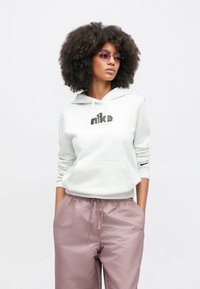 Hellgrauer Hoodie mit schwarzem Logo, Kängurutasche und Kordelzug; kombiniert mit pinken Sporthosen mit Logo an der Seite.