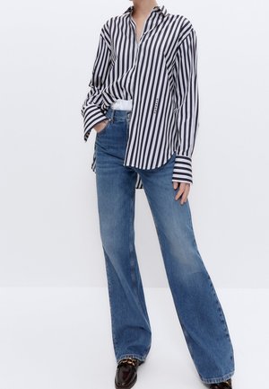 Personne portant une chemise à rayures verticales noir et blanc rentrée dans un jean bleu taille haute à jambes larges avec des mocassins noirs.
