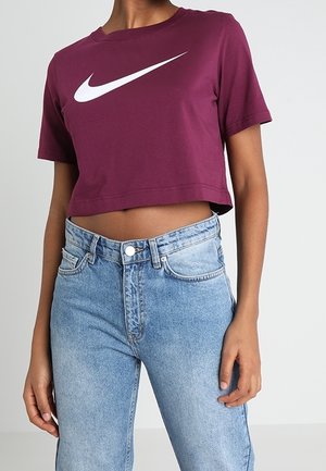 Osoba ubrana w krótki bordowy t-shirt Nike i jasnoniebieskie jeansy z wysokim stanem, stojąca z jedną ręką swobodnie opuszczoną wzdłuż ciała.