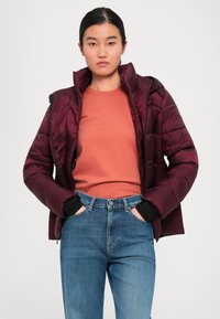 Young woman with hands in jean pockets wearing a maroon puffer jacket over an orange shirt against a plain background.