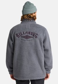 Grå fleece-jakke med en tekstureret overflade, høj krave og broderet "Billabong"-logo i lilla på ryggen.