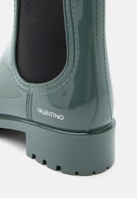 Bota de borracha verde brilhante com painel elástico preto, bico arredondado e sola robusta. Apresenta o logótipo "Valentino" em relevo perto do calcanhar.