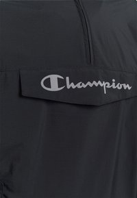 Schwarzer Stoff mit einer horizontalen Klapptasche, die das silberne "Champion"-Logo trägt, sowie einem Reißverschluss über der Tasche.