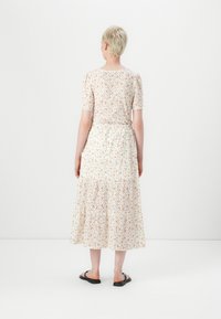 Robe midi fleurie avec des manches courtes, fond crème, motif floral orange et vert, dos smocké, jupe à volants, portée avec des sandales noires.