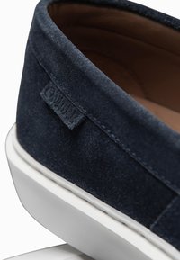 Gros plan du talon d'une chaussure en daim bleu marine avec une semelle en caoutchouc blanche et une petite étiquette de marque en tissu sur le côté.