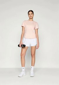 Svetloružová kratkosrstá tričko s krátkymi rukávmi v kombinácii s bielymi športovými šortkami. Model drží dva čierne činky. Jednoduchý, priliehavý dizajn, hladká textura.