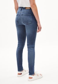 Mörkblå skinny jeans med hög midja, med två bakfickor och en etikett på midjebandet. Bärs med ljusa sandaler.