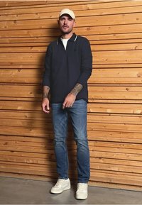 Námořnický polo s dlouhým rukávem a bílým límcem, modré džíny a bílé tenisky. Model stojí proti dřevěné zdi s vodorovnými prkny.