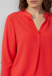 Rode blouse met lange mouwen en een notch-kragen, gemaakt van een textuurstof. Bevat opgerolde mouwen met knoopaccenten.
