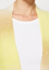 Primer plano de una persona que lleva una camiseta de rayas blanca debajo de un cárdigan de punto amarillo y beige.