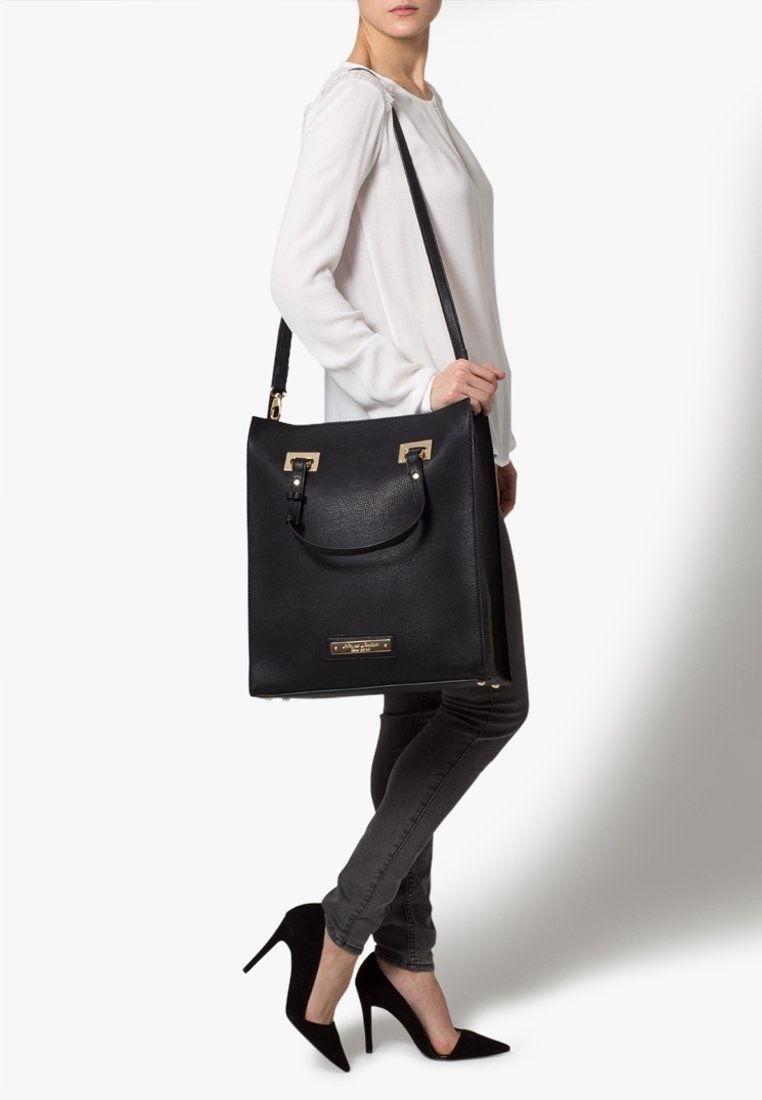 Sac fourre-tout noir, structuré, avec une finition texturée, des accents en métal doré et une poignée supérieure. Porté avec une bandoulière.
