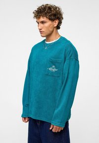 Teal sweatshirt met lange mouwen, voorzien van een borstzak met geborduurd logo, ontspannen pasvorm en zachte textuur.