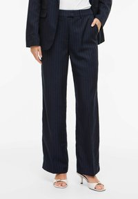 Femme portant un pantalon large bleu marine à fines rayures avec un blazer assorti et des sandales blanches à talons, main dans la poche.