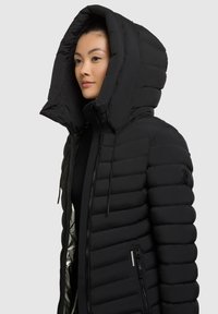 Giacca a vento nera con grande cappuccio, design trapuntato e tasche laterali con zip. Presenta una texture liscia ed è realizzata in materiale leggero.