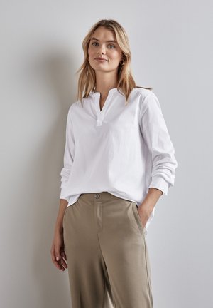 Blouse blanche à manches longues avec un col mandarin, présentant une coupe ample et une texture douce, associée à un pantalon taille haute taupe.
