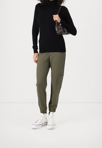 Black turtleneck sweater, olive green jogger pants with elastic cuffs, and white high-top sneakers. Model holds a small brown bag.