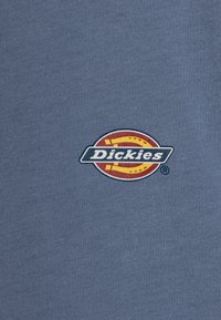 Szürke pamut póló, amelyen egy kör alakú logó található a "Dickies" felirattal fehér színben, piros és sárga kiegészítőkkel körítve.
