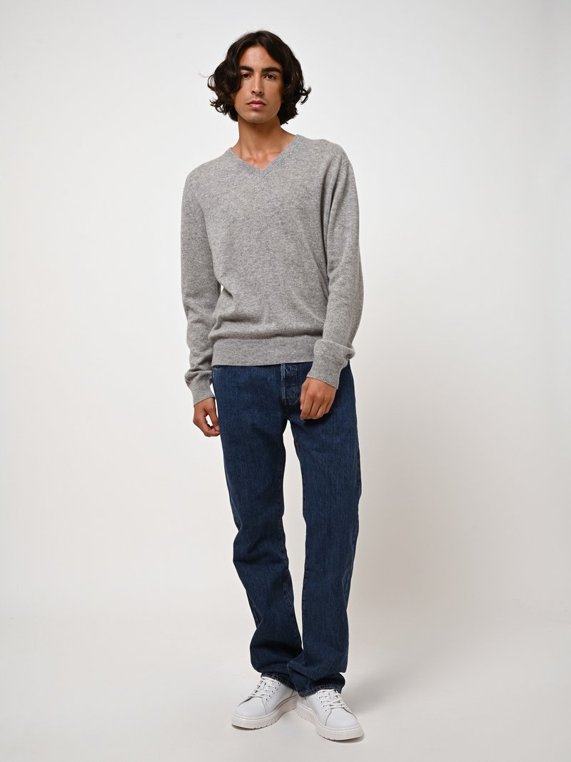 Grauer Strickpullover mit V-Ausschnitt, kombiniert mit dunkelblauen Jeans. Das Model trägt weiße Sneakers. Weicher Stoff, minimalistisches Design und klassischer Schnitt.