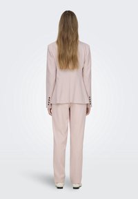 Costume sur mesure rose clair comprenant un blazer structuré avec des boutons en accent et un pantalon assorti, présenté de dos.