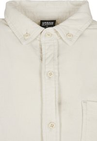 White corduroy shirt featuring a button-up design, pointed collar, front pocket, and plastic buttons. Textured fabric with ribbed pattern.