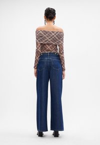 Top de mangas longas e ombros à mostra em padrão de plaid transparente, combinado com jeans de denim azul de cintura alta e perna larga. Textura do tecido suave e silhueta.