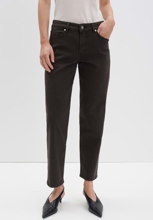 Femme portant un jean noir slim et des chaussures noires à talons bas et bouts pointus, debout avec une main dans la poche sur un fond uni.