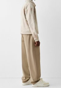 Beige mokkanahkatakki leveillä hihoilla, yhdistettynä löysiin beigeihin housuihin ja valkoisiin tennareihin. Asussa on minimalistisia design-elementtejä.