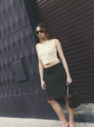 Giovane donna con occhiali da sole, top beige senza maniche, gonna nera a pois e sandali, che tiene una borsa scura, in piedi accanto a una porta metallica con saracinesca.