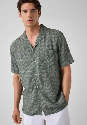 Jeune homme portant une chemise verte à manches courtes boutonnée avec "GUESS" imprimé en continu et un pantalon blanc sur un fond uni.