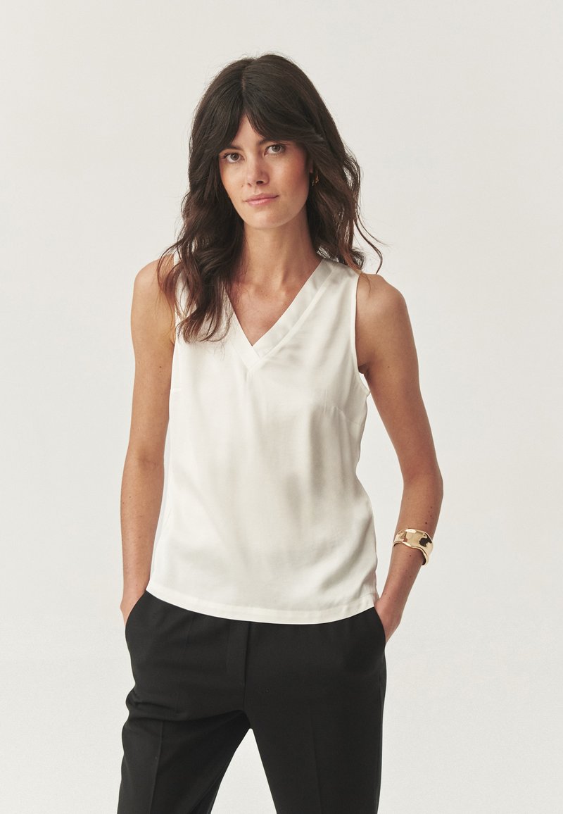 Blouse sans manches blanche avec un design en V, fabriquée en tissu lisse. Positionnée avec les mains dans les poches, associée à un pantalon noir.