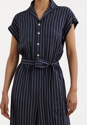 Woman wearing navy blue jumpsuit with white vertical stripes, short sleeves, button front, chest pocket, and tied waist belt.