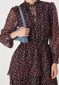 Námořnické květinové šaty s dlouhými, průhlednými rukávy, řaseným límcem a staženým pasem. Drží malou, světle modrou kabelku s texturovaným povrchem.