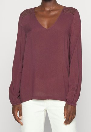 Bluzka w ciemnym kolorze mauve z długim rękawem, koronkowym detalem na ramionach, dekoltem w serek i marszczonymi mankietami, noszona z białymi spodniami.